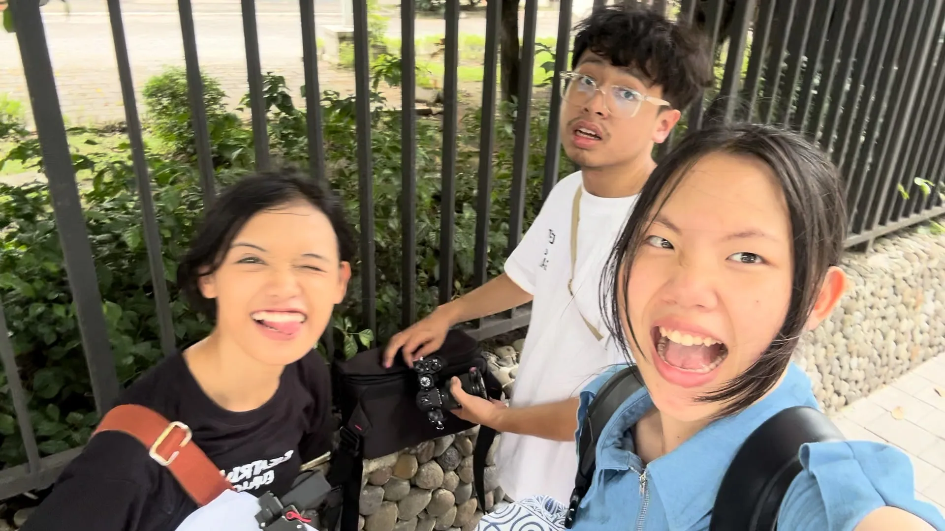 Behind the scenes, three team members pull faces for a selfie by a black iron fence, one of them holding a camera.